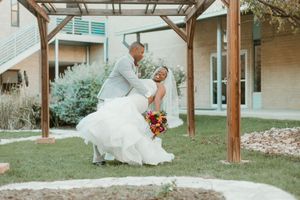 Groom dips bride in white wedding gown outdoors under wooden pergola in romantic wedding portrait.