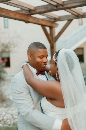 African American bride and groom share a tender embrace outdoors, groom in light gray suit with burgundy bow tie, bride in white veil.