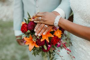 Bride and groom display wedding rings while holding a vibrant bouquet of orange lilies and red flowers.