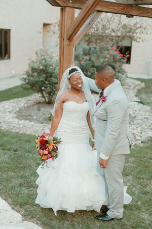 A smiling Black bride in a strapless lace gown holds a vibrant bouquet while standing with her groom outdoors.