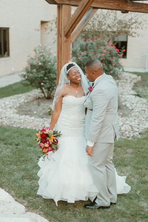 Smiling African American bride in strapless mermaid gown holds vibrant bouquet while groom in gray suit gazes at her outdoors.