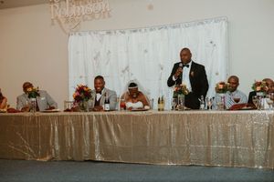 Wedding party seated at gold sequin head table during reception while groomsman delivers speech.