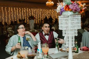 Two male wedding guests laugh and react animatedly at a reception table with drinks and floral centerpiece in background.