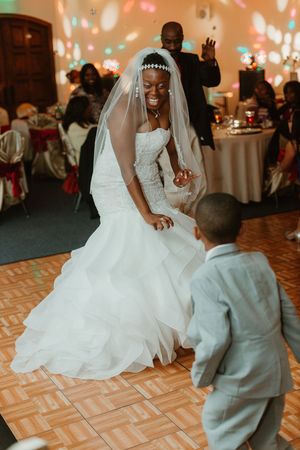 Joyful African American bride in white mermaid gown and veil dances with a young boy in gray suit on wedding reception dance floor.