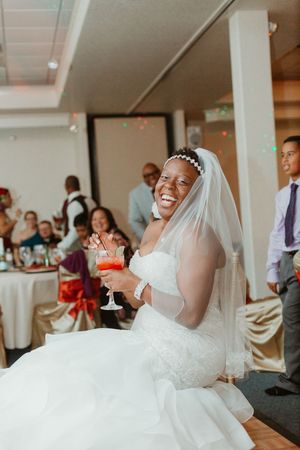 Radiant bride in white lace gown and veil smiling while holding a cocktail at wedding reception.
