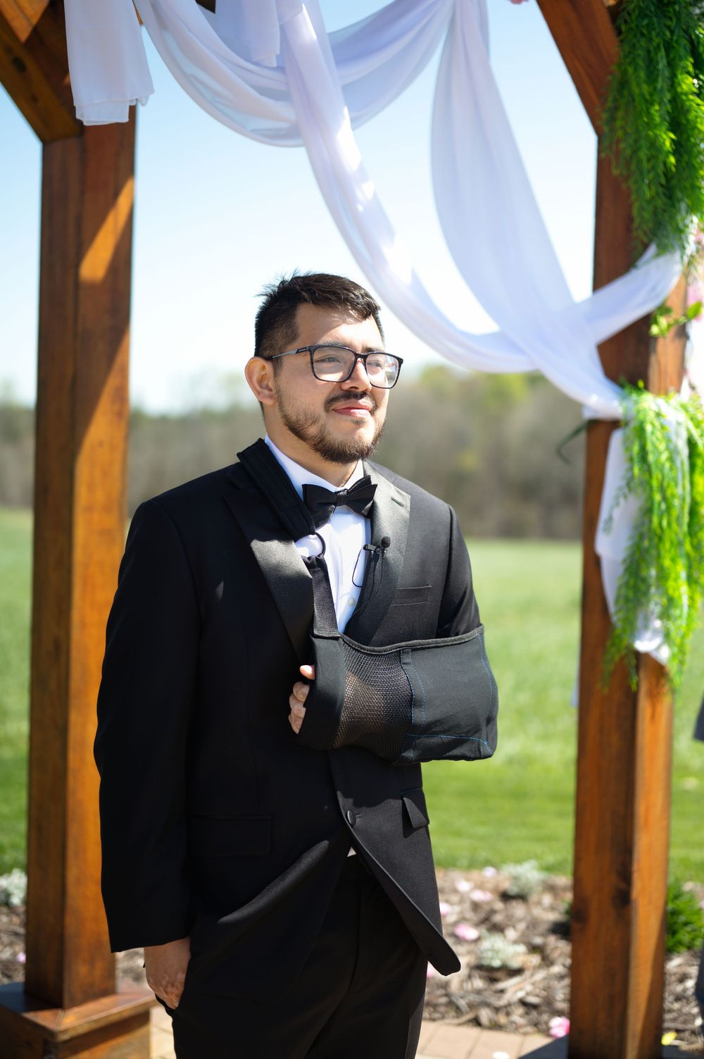 Groomsman in black tuxedo with arm sling stands under decorated wooden pergola at outdoor wedding ceremony.