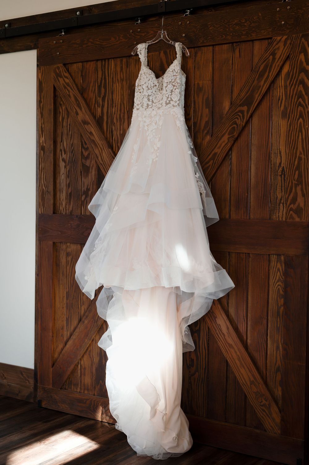 Elegant blush wedding gown with lace bodice hanging on rustic barn door.