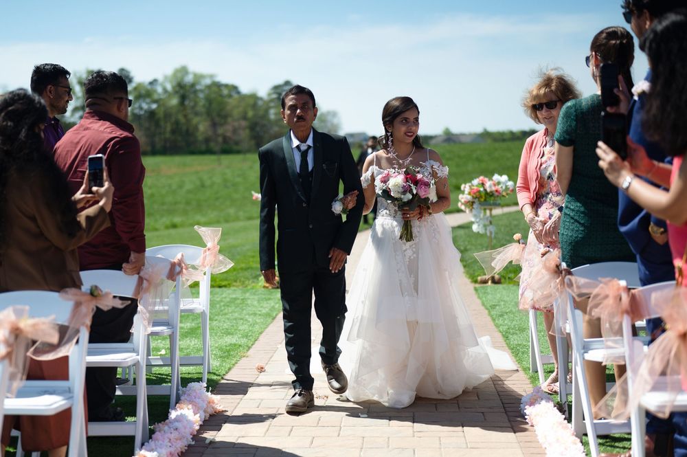Bride and groom walk down outdoor wedding aisle, guests cheering, green fields in sunny background.