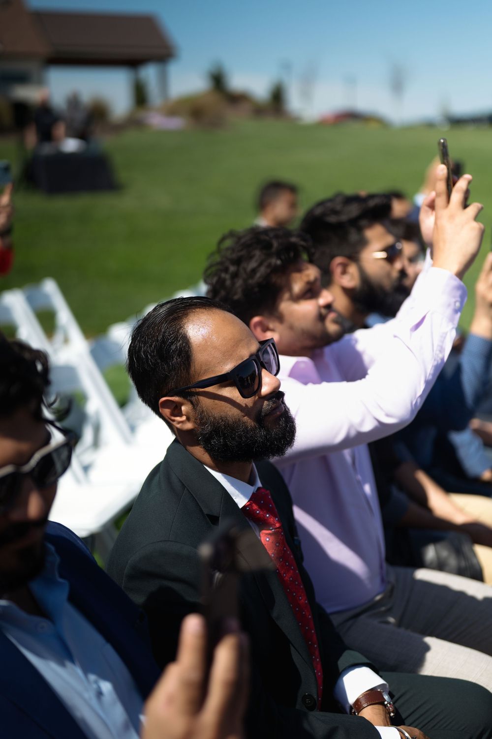 Wedding guest in sunglasses and red tie seated at outdoor ceremony.