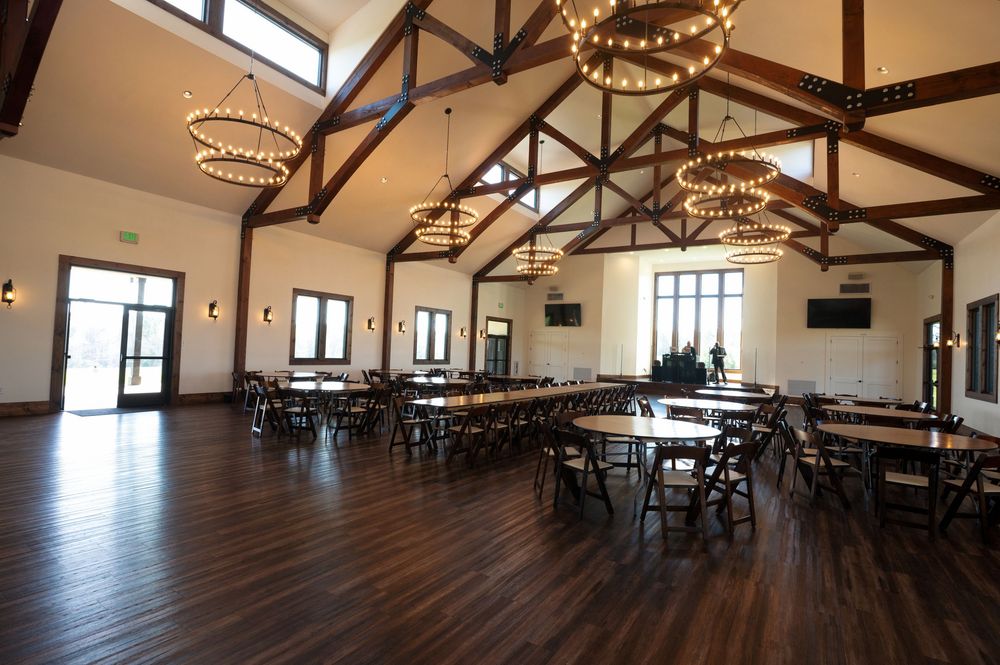 Elegant event hall with vaulted ceilings, wooden beams, circular chandeliers, and dark hardwood floors set with tables and chairs.