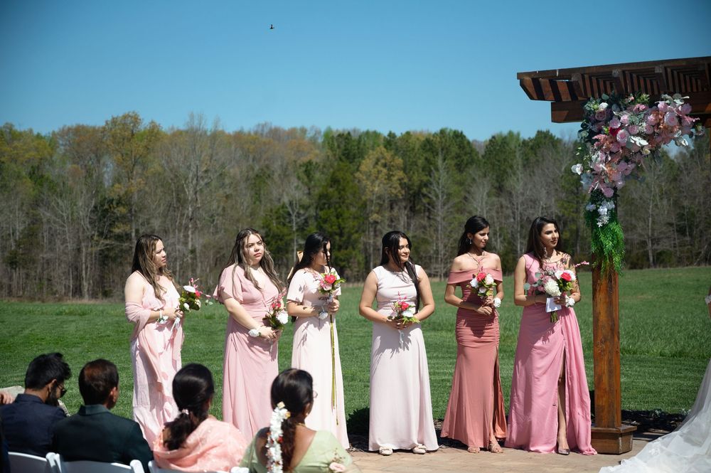 Six bridesmaids in pink dresses holding bouquets stand at outdoor wedding ceremony with wooded backdrop.