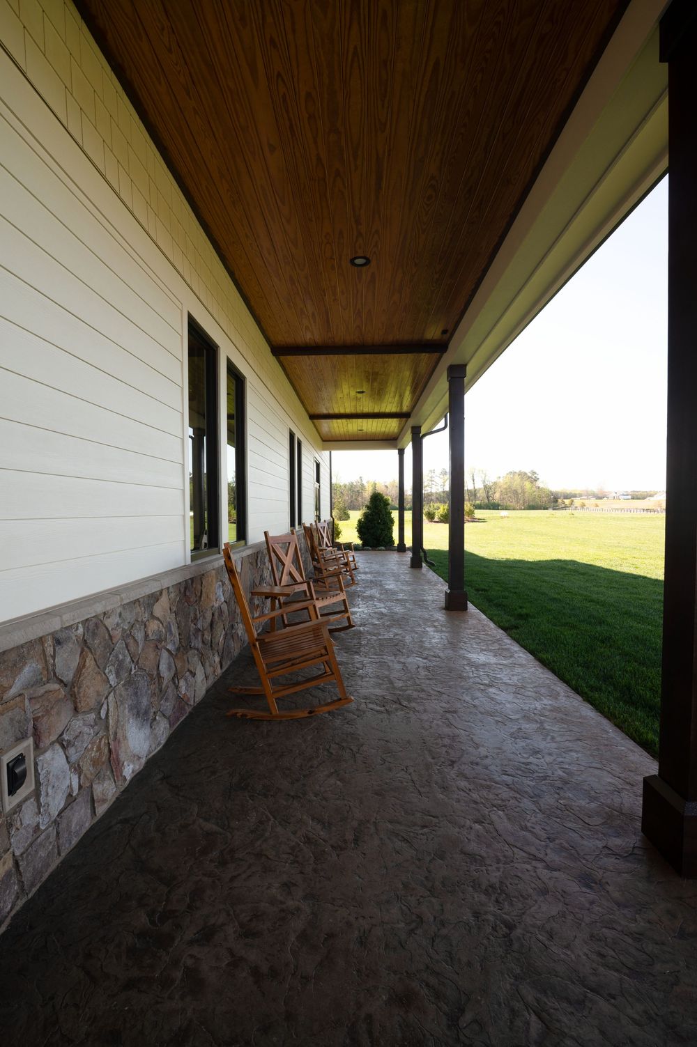 Spacious covered farmhouse porch with wooden rocking chairs, stone foundation, wood ceiling, and scenic green lawn views.