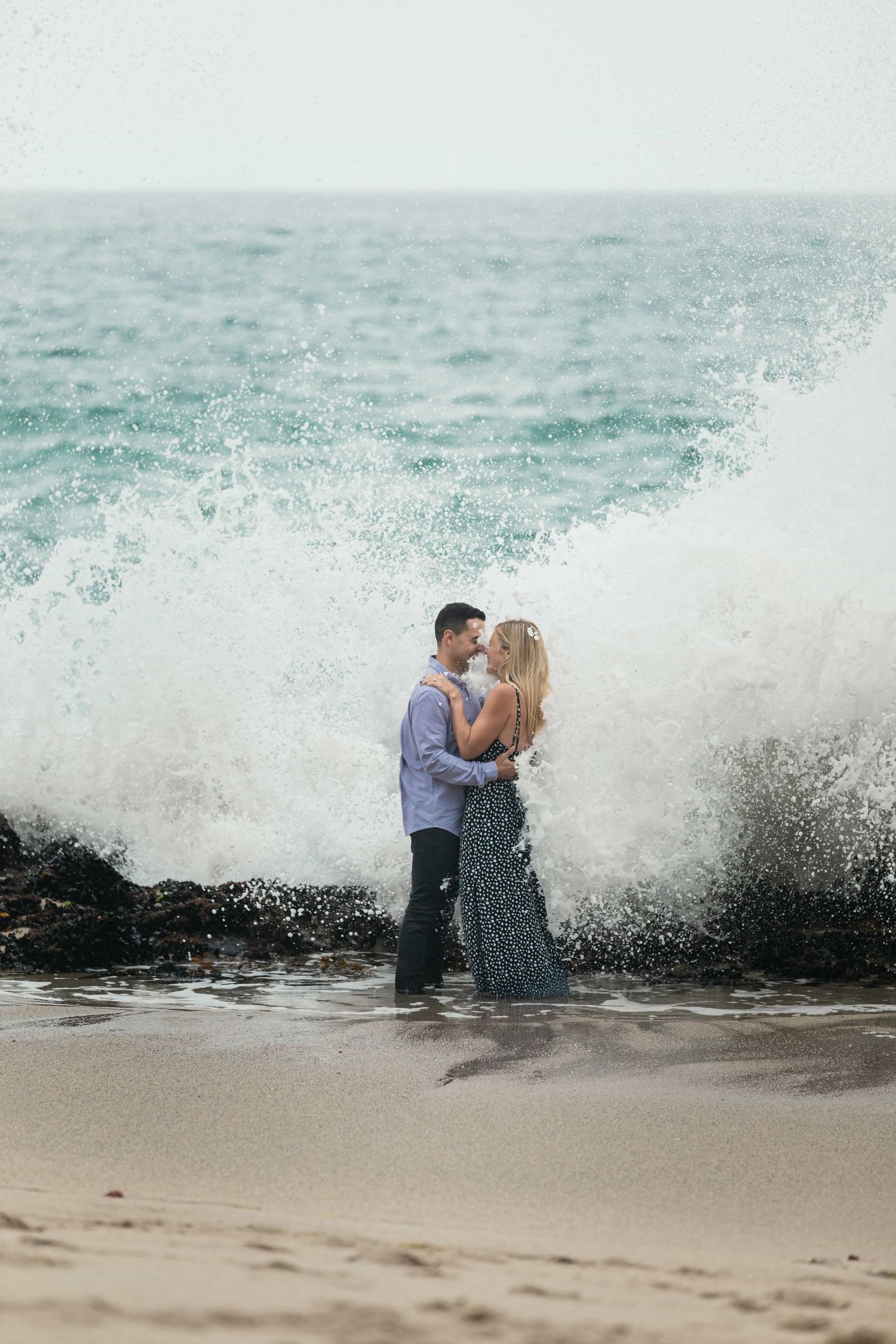 Couple embracing as a dramatic ocean wave crashes behind them on the beach.