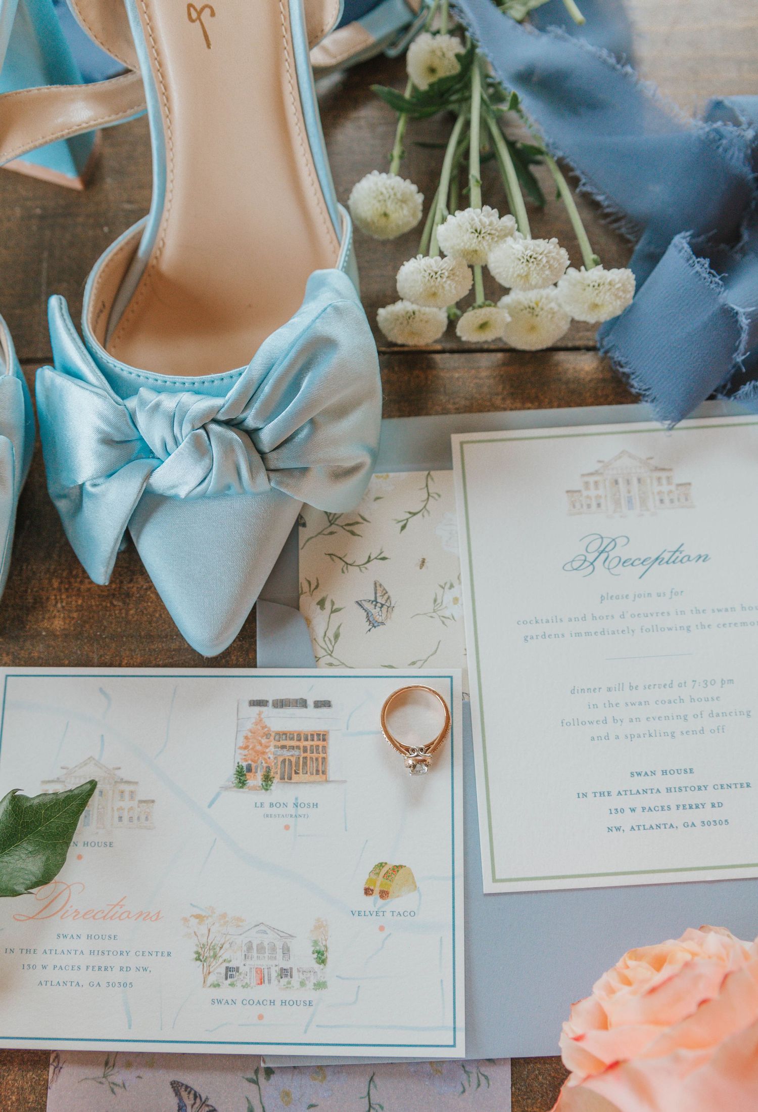 Light blue satin bow heels displayed with wedding reception invitations, a diamond ring, and white flowers on a rustic surface.