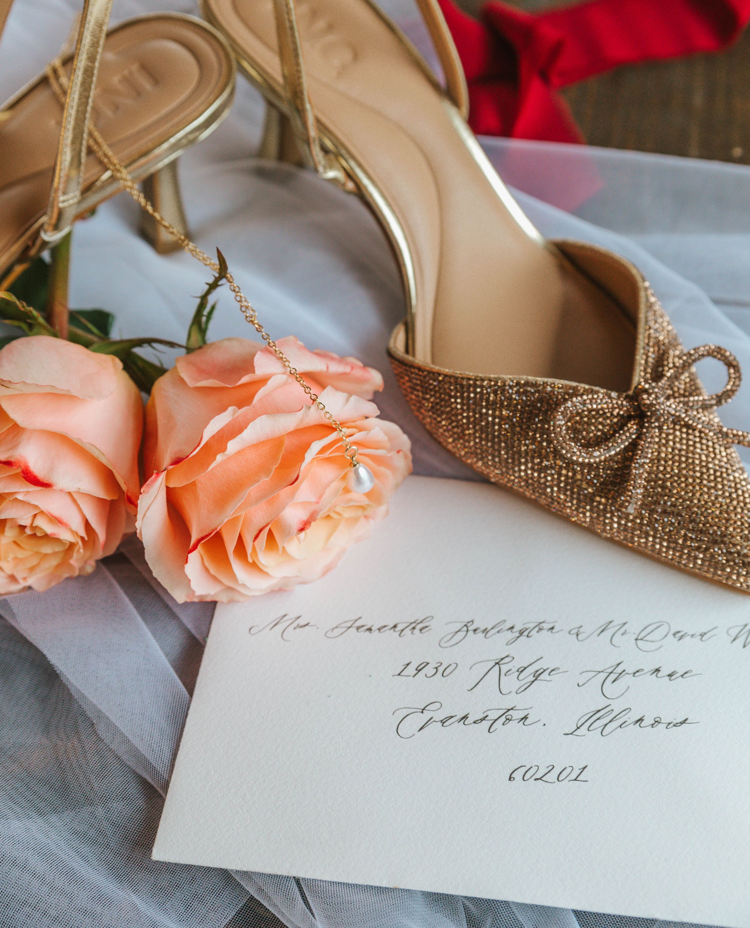 Gold glitter bow heels beside peach roses and elegant calligraphy wedding envelope on soft white fabric.
