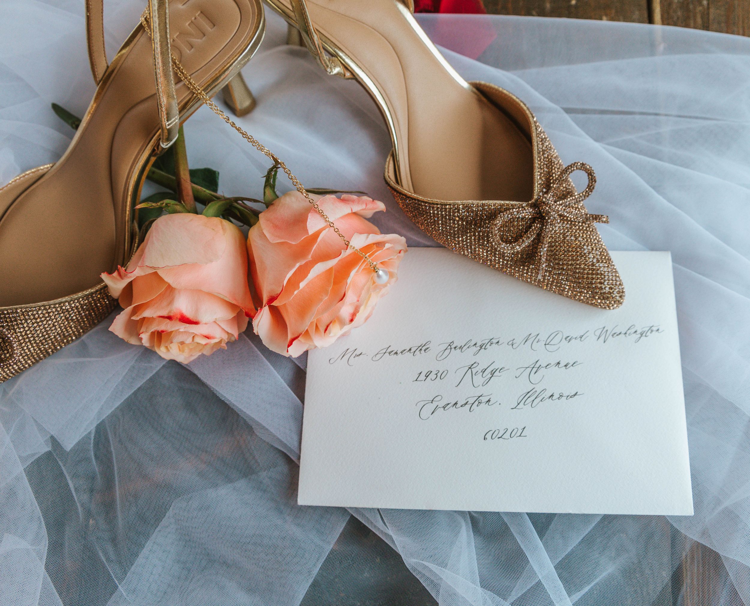 Gold bridal heels with burlap bows, peach roses, and a handwritten wedding envelope on white tulle fabric.