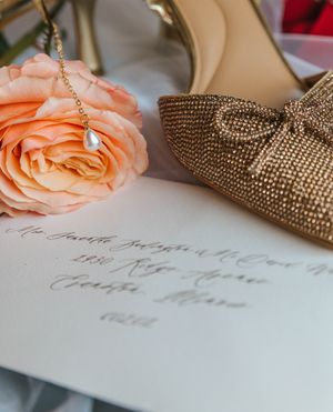 Gold beaded bow heels beside a peach rose, pearl necklace, and handwritten calligraphy envelope on white surface.