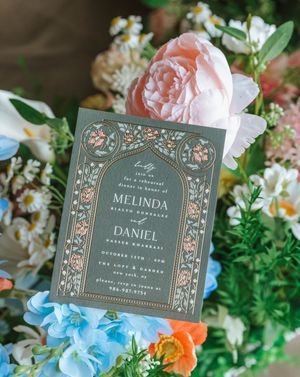 Elegant wedding rehearsal dinner invitation with floral arch design on sage green card, surrounded by pink and blue flowers.
