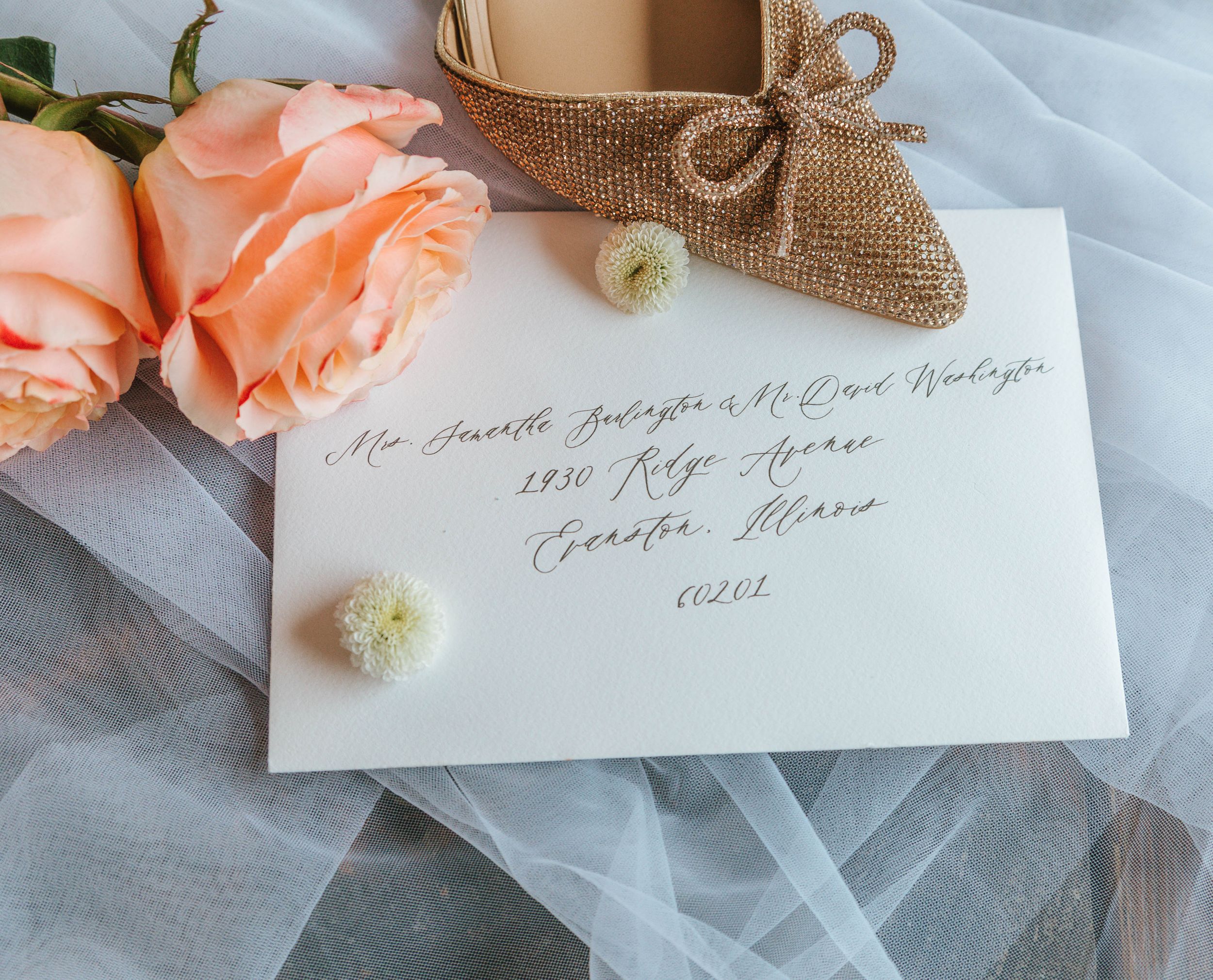 Elegant wedding envelope with gold calligraphy, peach roses, burlap bow, and white flowers on sheer tulle fabric.