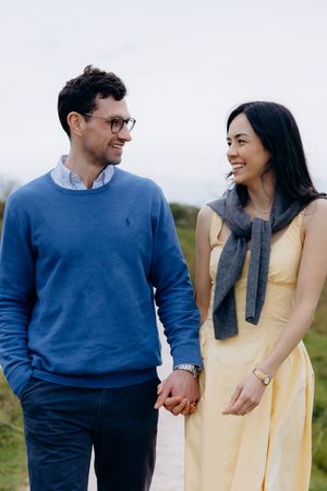 A smiling couple holds hands outdoors, man in blue sweater and glasses, woman in yellow dress with gray scarf.