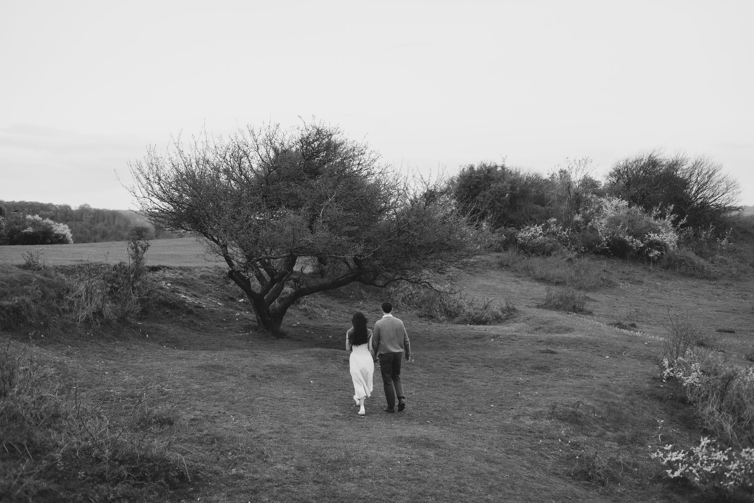 Black and white photo of a couple holding hands walking along a rural path surrounded by bare trees and open fields.