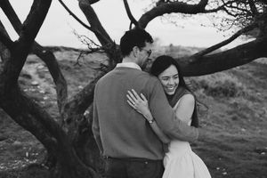 Black and white photo of a couple embracing and laughing near a gnarled tree in a scenic outdoor setting.
