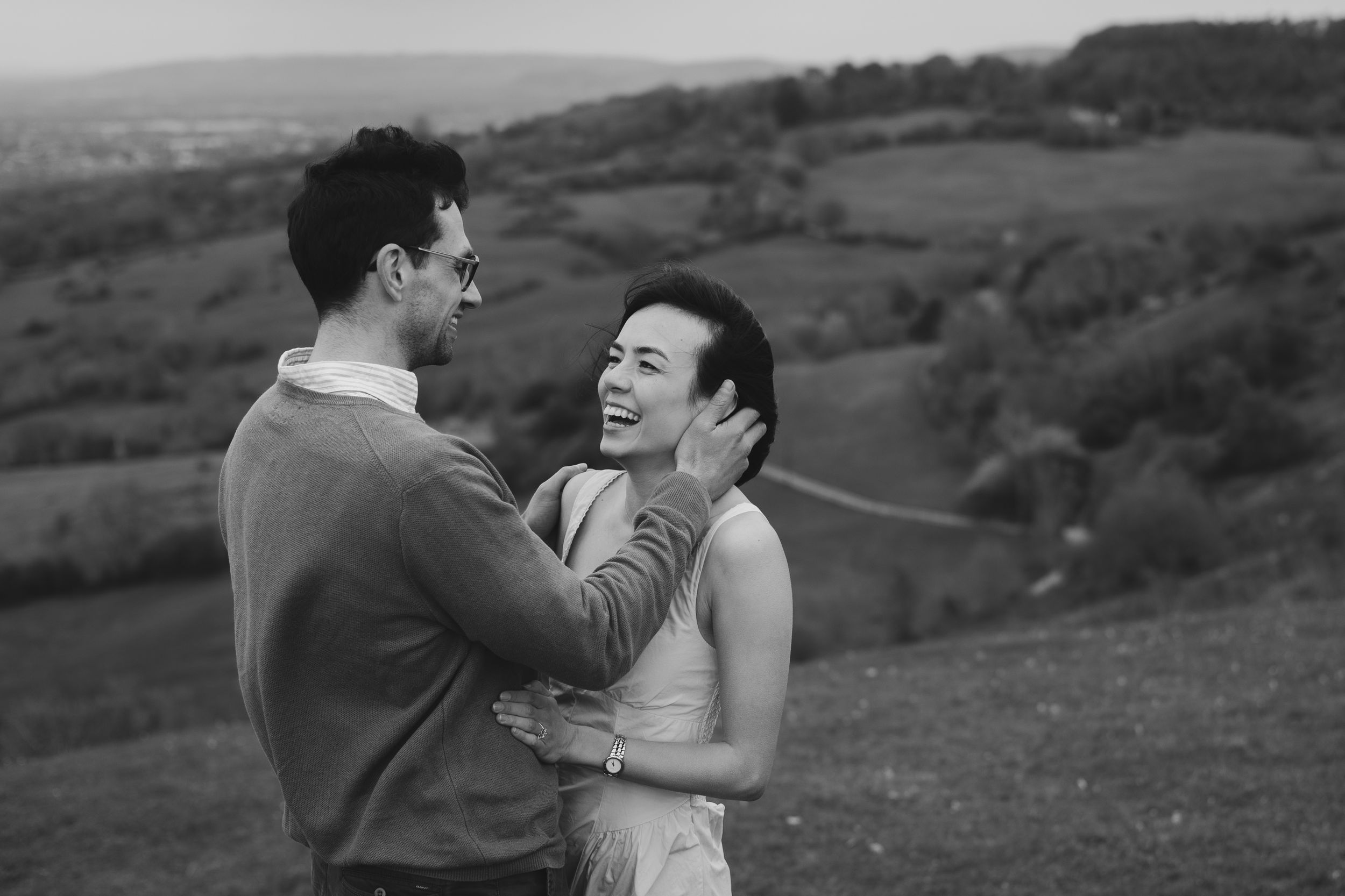Black and white photo of a laughing couple embracing on a scenic hillside with rolling green countryside in the background.