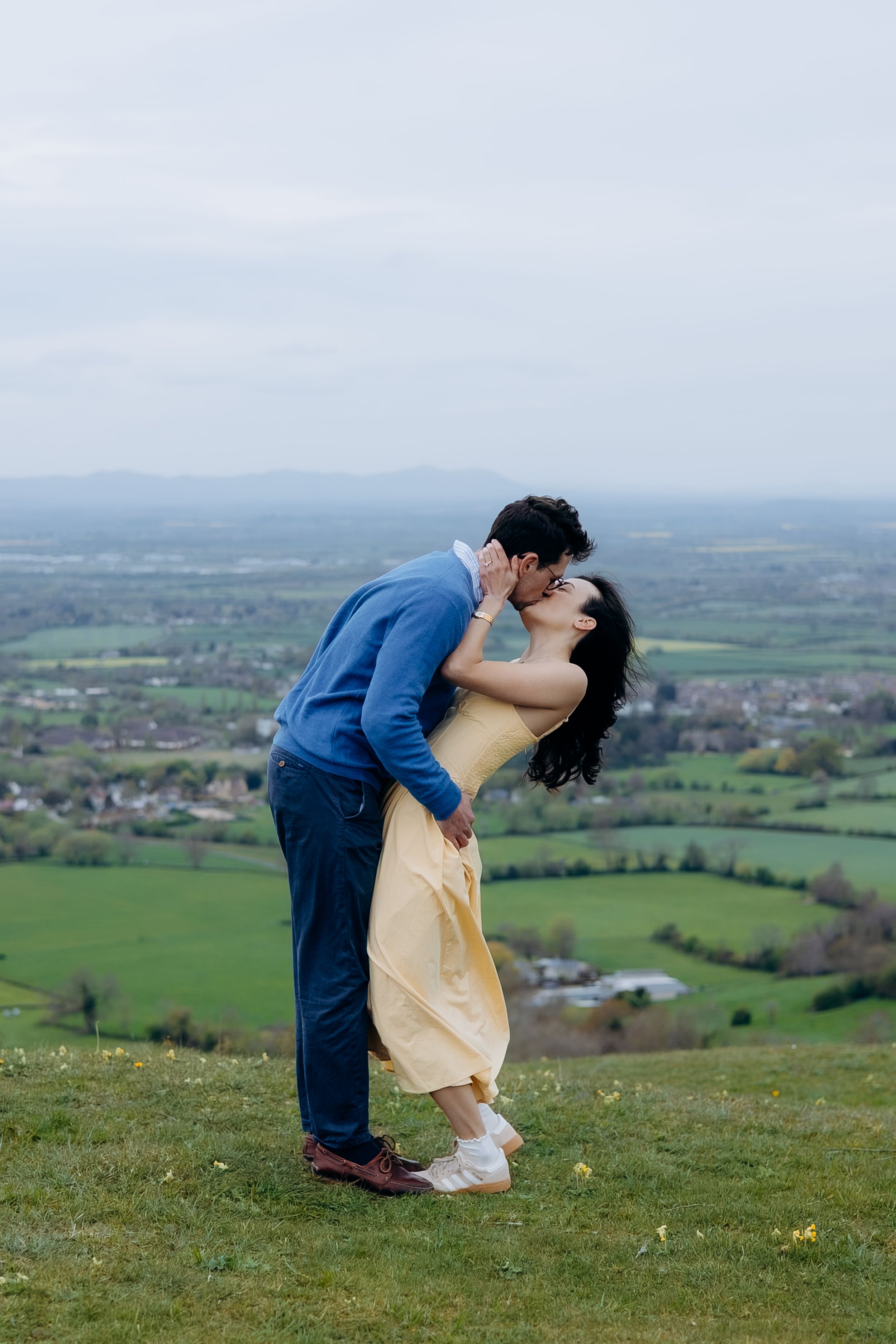 A couple shares a romantic kiss on a hilltop overlooking lush green countryside fields under an overcast sky.