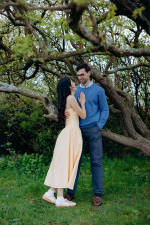Couple sharing a romantic moment under twisted tree branches in a lush green garden, woman in yellow dress, man in blue sweater.