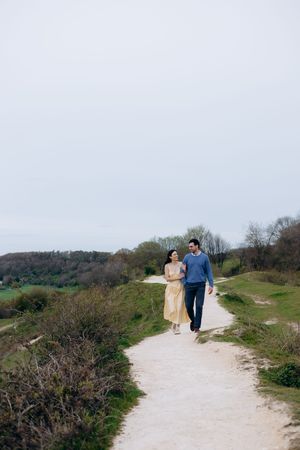 Couple walking hand in hand on a scenic countryside path surrounded by green hills and trees on an overcast day.