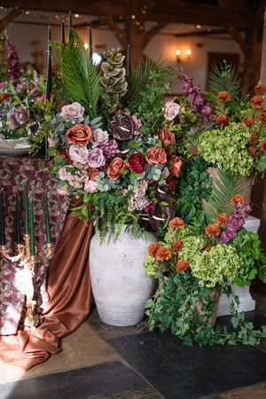 Large white vase overflowing with lush roses, hydrangeas, and tropical foliage in rich burgundy, coral, and green tones.