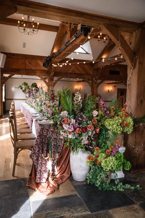 Rustic barn wedding reception with long floral table runner featuring lush greenery, roses, and hydrangeas under string lights.