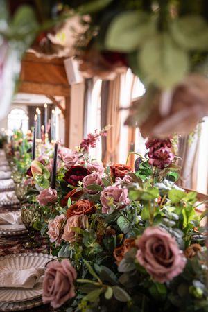 Elegant wedding table centerpiece with dusty pink roses, deep red blooms, greenery, and tall black taper candles.