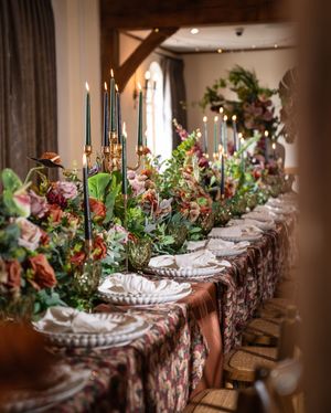Elegant wedding reception table with lush floral garland, dark taper candles, and ornate place settings in a rustic venue.