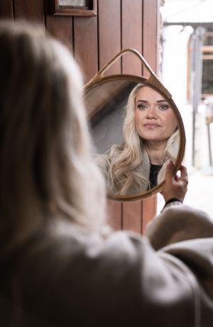 Blonde woman holding a teardrop-shaped rattan mirror, smiling at her reflection outdoors.