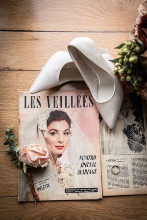 White bridal heels resting on a vintage French Les Veillées wedding magazine with a rose and gold rings on wood.