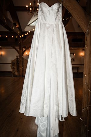 A strapless white ball gown wedding dress hanging in a rustic barn venue with warm Edison bulb lighting.