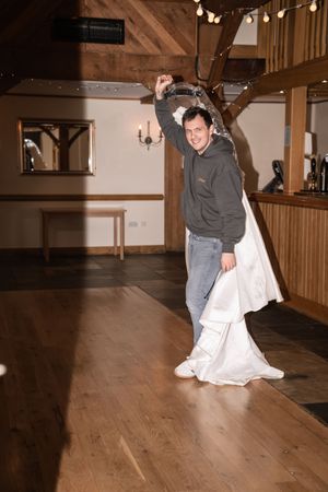 Man in hoodie twirling a white wedding dress on a wooden dance floor in a rustic barn venue.