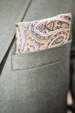 A paisley pocket square in gold and blue tucked into a sage green suit jacket breast pocket.