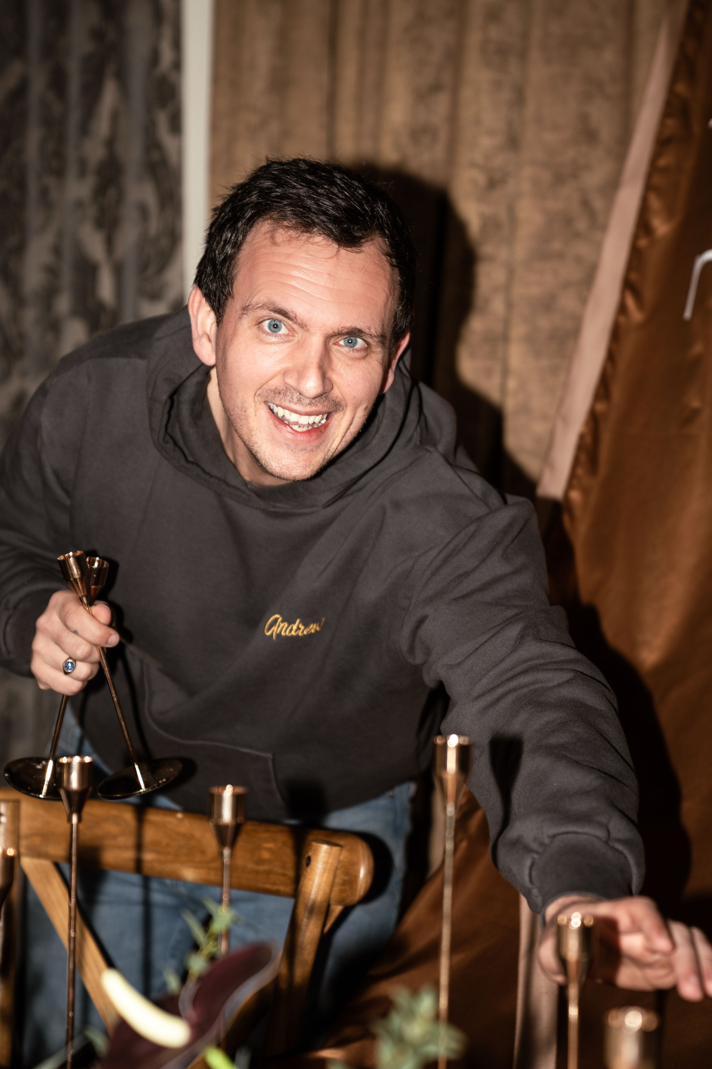 Smiling man in gray hoodie leaning over table with candles and decorative items in dimly lit room.