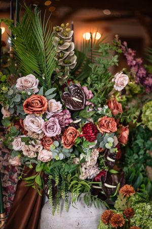 Elegant floral arrangement featuring coral and mauve roses, dark anthuriums, and eucalyptus in a rustic stone vase.