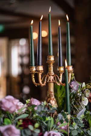 Gold ornate candelabra with five lit dark green taper candles surrounded by pink and burgundy floral arrangements.