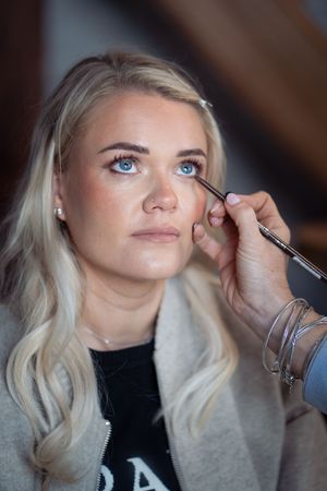 Makeup artist applying eyeshadow with a brush to a blonde blue-eyed woman wearing a gray coat.