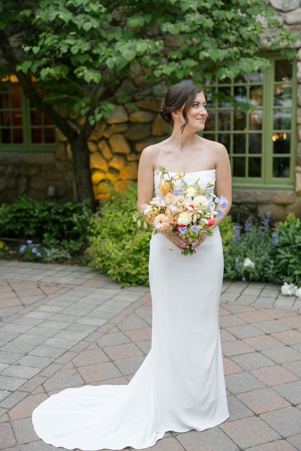 Bride in strapless white gown holding colorful bouquet stands on brick patio surrounded by lush garden greenery.