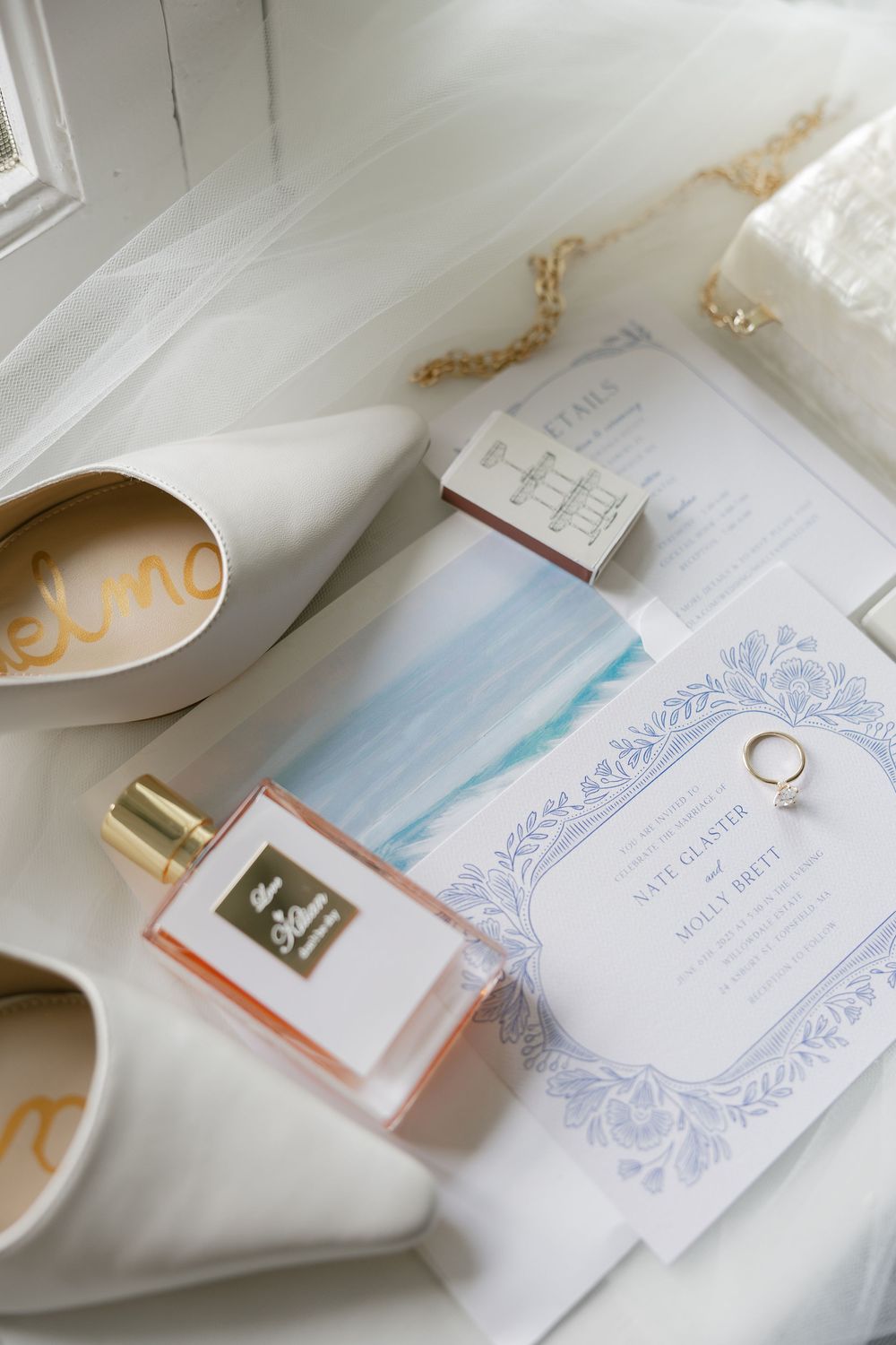Elegant wedding flat lay featuring white bridal heels, perfume bottle, ring, and blue watercolor invitation suite.