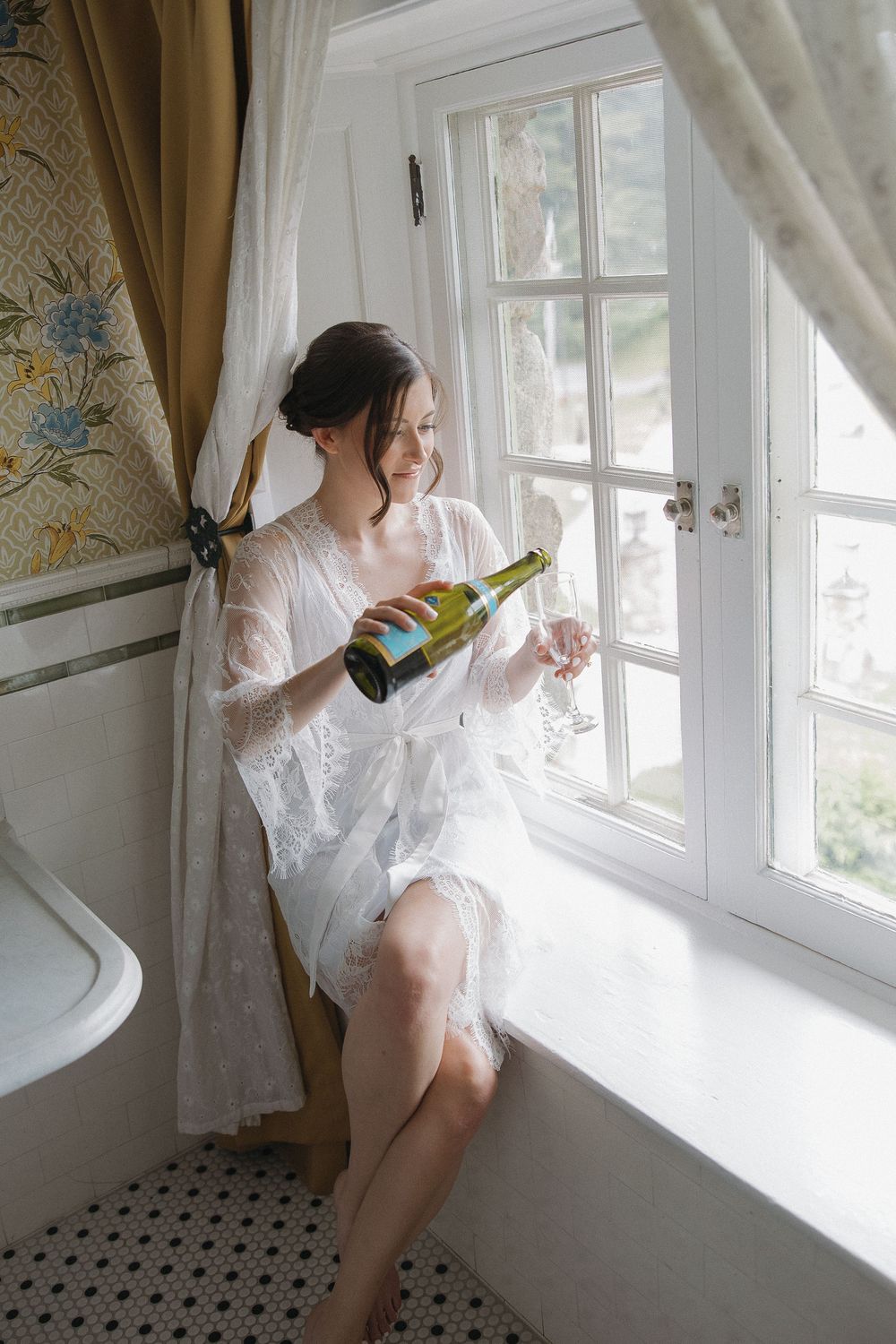 Bride in white lace robe pouring champagne by a sunlit window, elegant bridal morning preparation.