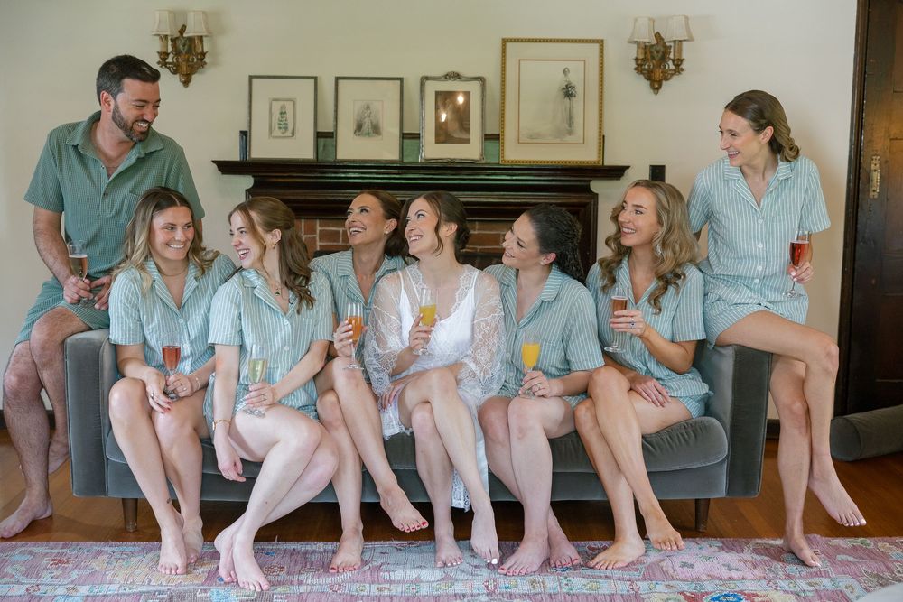 Bridesmaids in matching mint pajama sets laughing together on a gray sofa during a bridal getting-ready party.