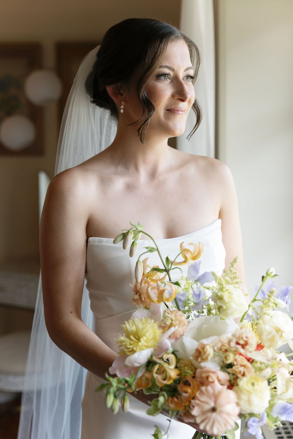 Elegant bride in strapless white gown holding colorful peach and pastel wedding bouquet with veil, gazing out window.