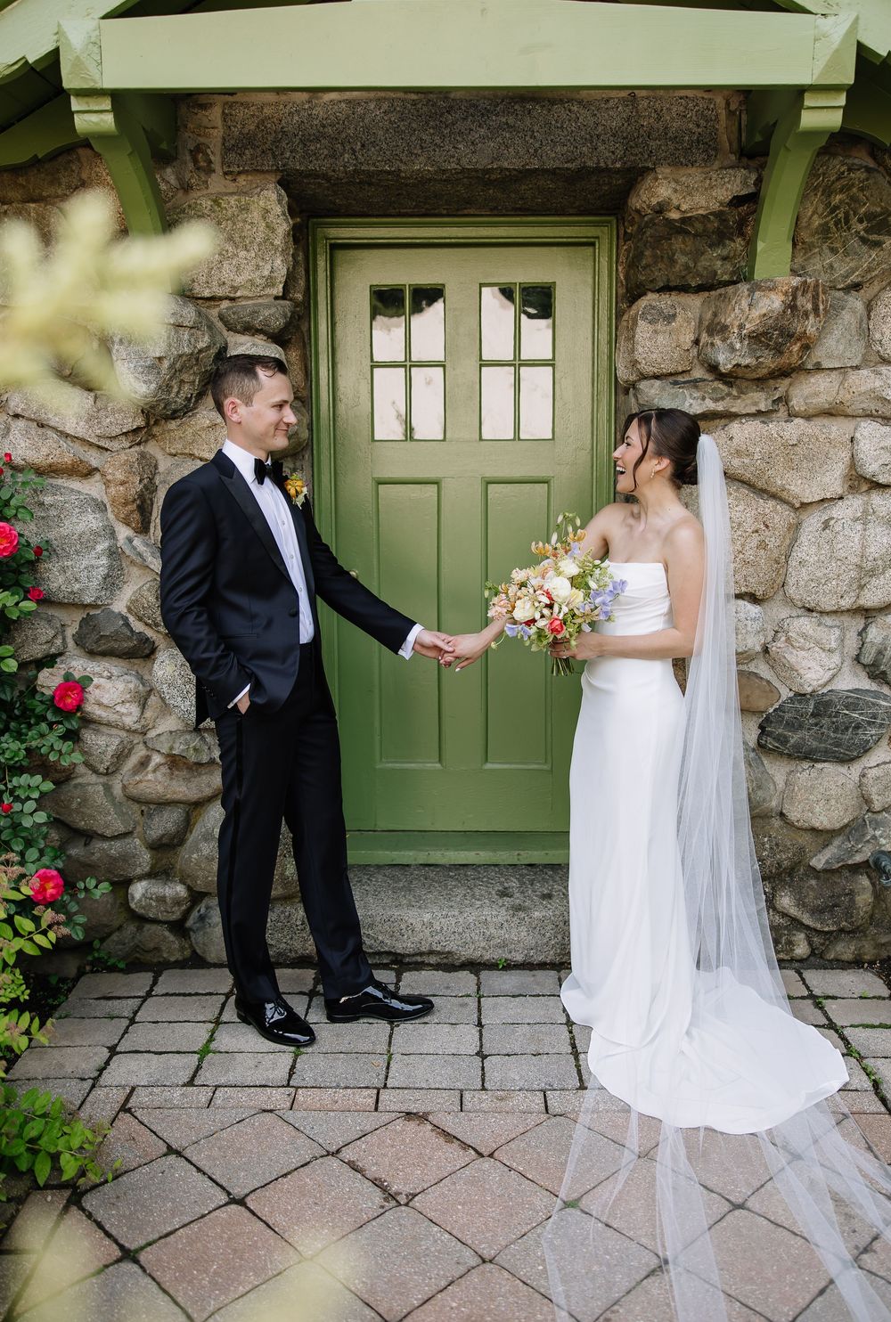 Bride and groom hold hands facing each other before a green door of a stone building on wedding day.