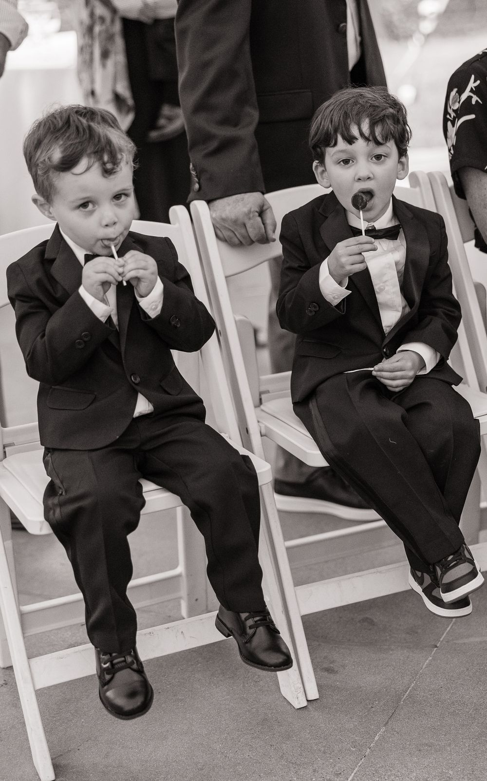 Two young boys in black suits sit on white chairs eating lollipops during a wedding ceremony.